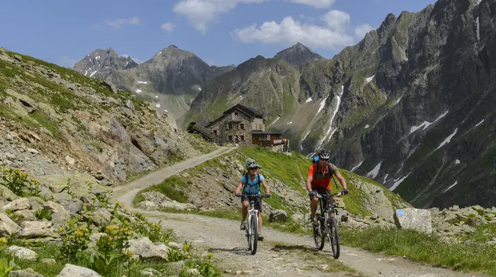 Zwei Mountainbiker*innen fahren auf einem Schotterweg in den Bergen. Im Hintergrund eine Hütte. | © DAV/Wolfgang Ehn